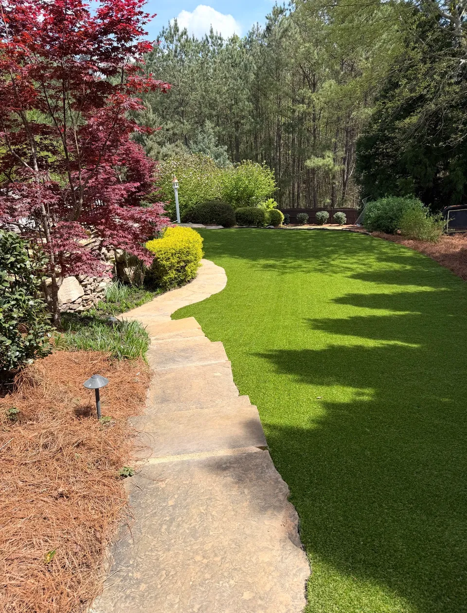Artificial turf bordered by a stone path, pine straw, and established plantings